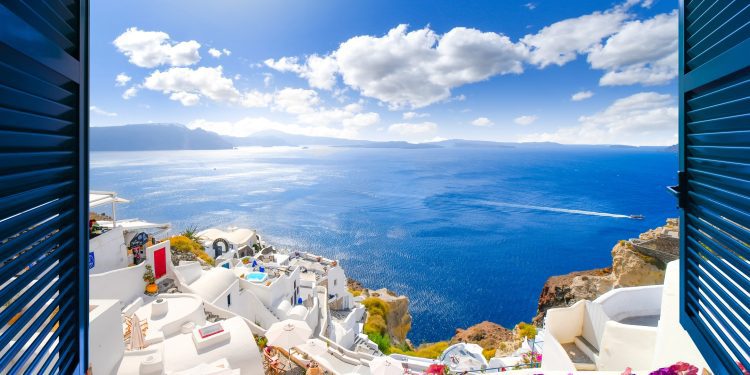 View through an open window with shutters of the whitewashed village of Oia rising above the blue Aegean Sea and the caldera on the island of Santorini, Greece.