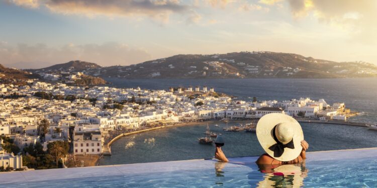 A woman with a glas of wine in a swimming pool enjoys the view over the town of Mykonos island, Greece