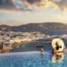 A woman with a glas of wine in a swimming pool enjoys the view over the town of Mykonos island, Greece