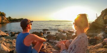 Sithonia - Rear view of happy couple admiring the sunset on a remote pebble beach on the peninsula of Sithonia, Greece